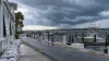 Barcos atracados numa marina na costa do Golfo sob nuvens escuras de tempestade, com sacos de areia a revestir os edifícios à beira-mar.