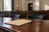 An unmarked case folder and reading glasses on a desk in an empty government hearing room.