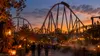 A large steel roller coaster above an autumn-decorated theme park midway at dusk in Southern California