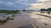 Floodwater covering a rural road and adjacent fields in southeast Nebraska beneath clearing storm clouds, shown as a restrained editorial illustration