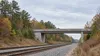 AI-generated editorial illustration of a generic railway corridor and road overpass in a wooded North Carolina setting; not a photograph of S-Line construction.
