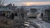 A humanitarian evacuation bus traveling on a damaged rural road near a checkpoint at dawn, shown as an illustrative editorial scene