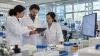 Three biopharmaceutical researchers review a tablet beside laboratory equipment and a project schedule.