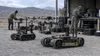 Technicians prepare uncrewed aerial drones and tracked ground robots at a generic military test range.