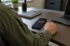 An anonymous person in a plain olive-drab uniform seated at a desk beside an unbranded smartphone displaying abstract market charts.
