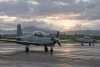 Two unmarked single-engine propeller training aircraft parked on a tropical airfield under overcast skies.