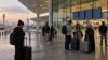 Travelers with luggage check phones outside a modern airport terminal at dawn during flight disruptions.
