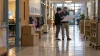 Dois funcionários da escola examinam uma planta não identificada num corredor tranquilo de uma escola primária, onde existem caixas de mudanças e carrinhos de livros.