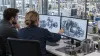 Dois engenheiros analisam um projeto mecânico tridimensional sem identificação em monitores acima de uma área de produção.