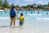 A child and supervising adult wear plain life jackets at the shallow edge of a generic water-park wave pool.