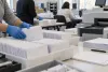Election workers in a county office organize sealed ballot envelopes beside processing equipment.
