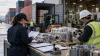 Workers review unbranded imported machine components beside shipping containers and factory equipment at an industrial loading dock.
