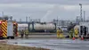Hazmat responders watch a venting intermodal tank from behind a safety perimeter at a generic industrial transport yard.