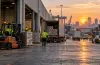 Workers and trucks move freight through a warehouse and port logistics hub at sunrise.
