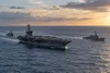 A generic aircraft carrier sailing with two escort ships on open ocean at sunset.