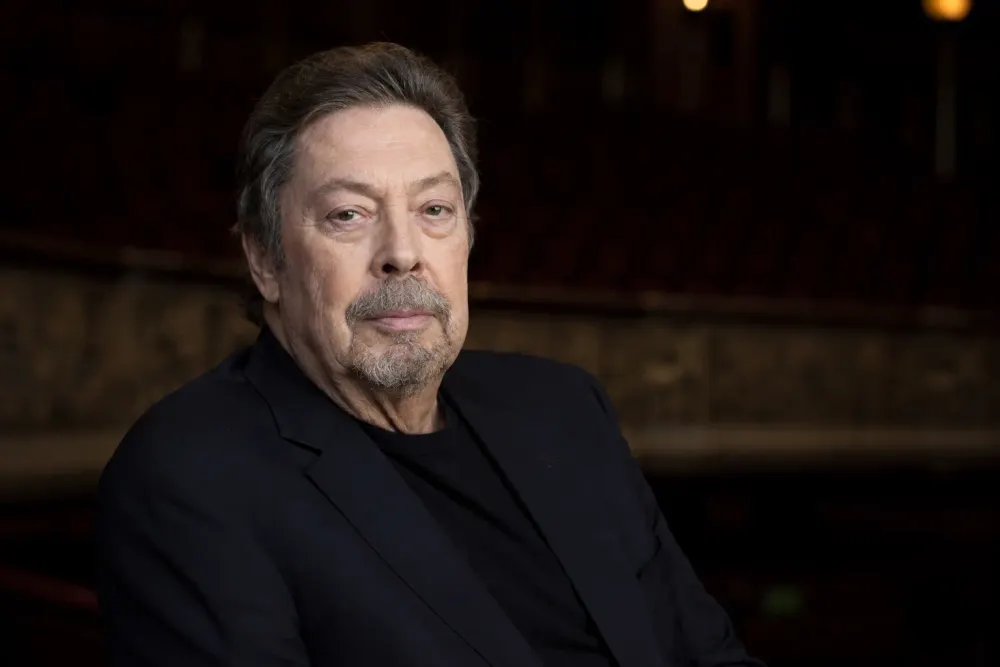 Photorealistic editorial portrait of actor Tim Curry in later life seated in a dark theater.
