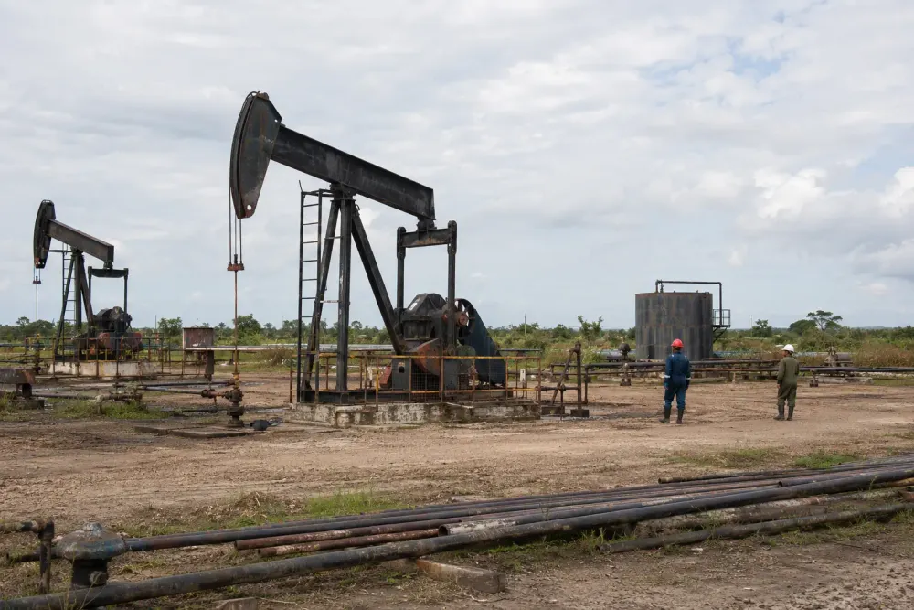 A weathered oil field with pumpjacks, pipes, storage equipment and two distant workers