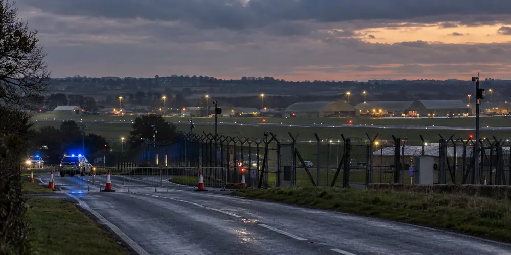 Illustrative dusk view of a secured road and perimeter fence beside a military airfield in rural England.
