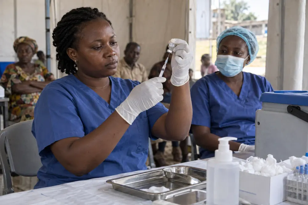 A health worker prepares an unlabeled vaccine dose inside a temporary clinic while a colleague waits nearby.