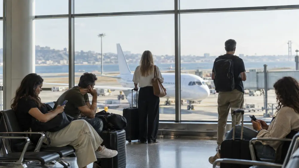 Travelers wait beside carry-on bags in a sunlit airport departure area overlooking an unbranded passenger jet.