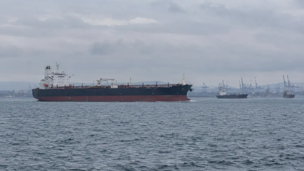 An unbranded crude oil tanker passes a commercial port under an overcast sky.
