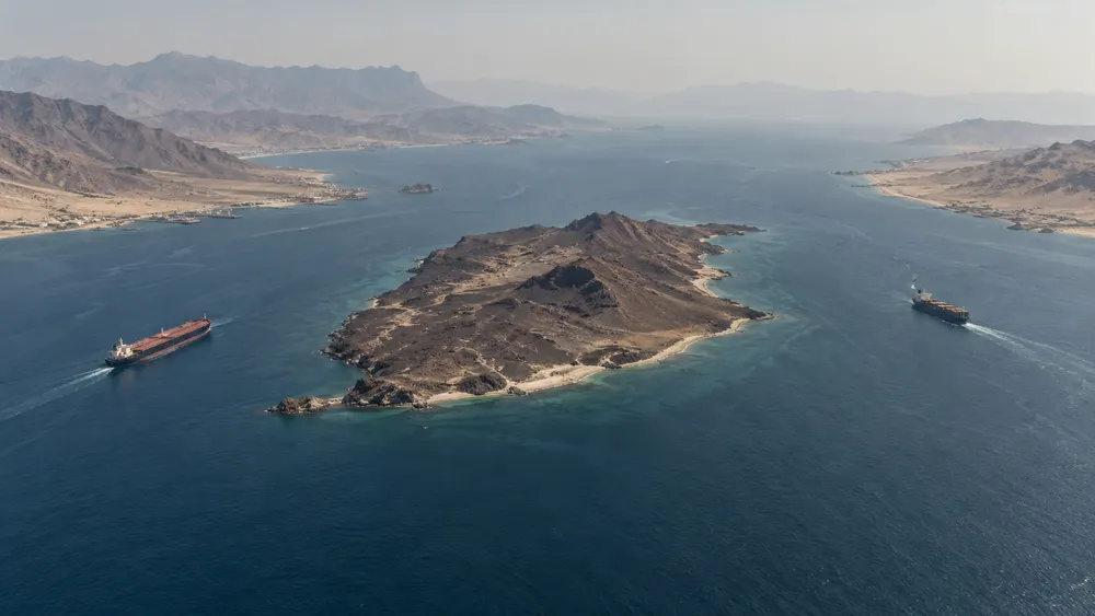 Aerial editorial view of a barren island dividing a narrow sea passage as an oil tanker and cargo ship transit on opposite sides.