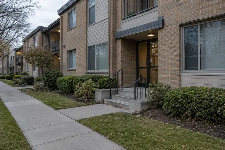 A modest apartment complex and quiet city sidewalk at dawn, representing federal homelessness housing programs.