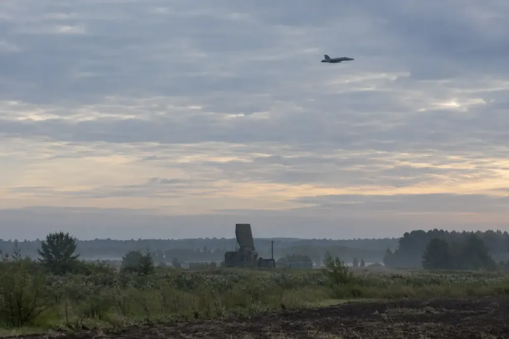 A distant fighter aircraft above a radar installation in a Baltic countryside landscape