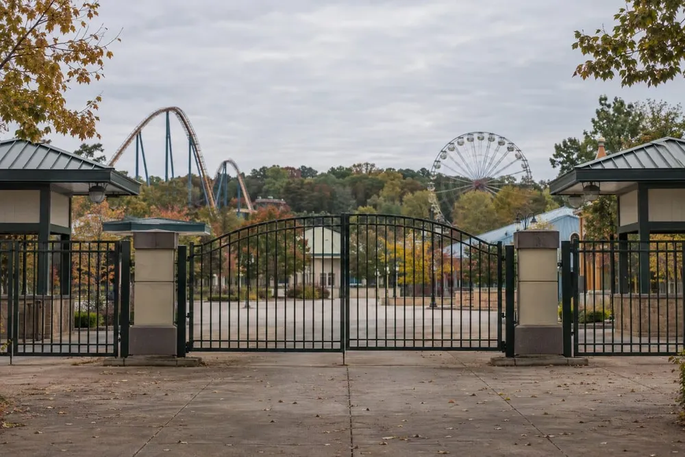 Editorial illustration of a quiet, unbranded amusement park entrance with idle coaster structures beyond the gates