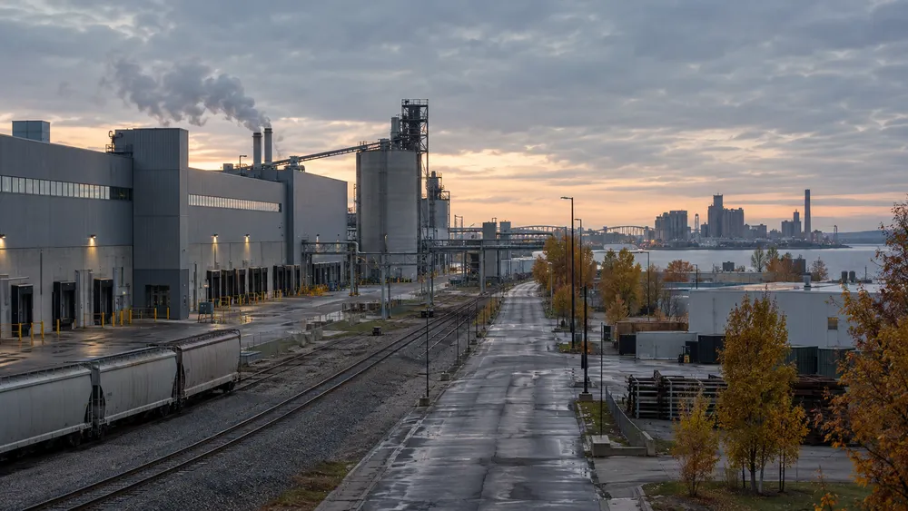 Quiet generic Michigan industrial corridor with factory buildings, rail lines and autumn trees beside a Great Lakes shoreline.