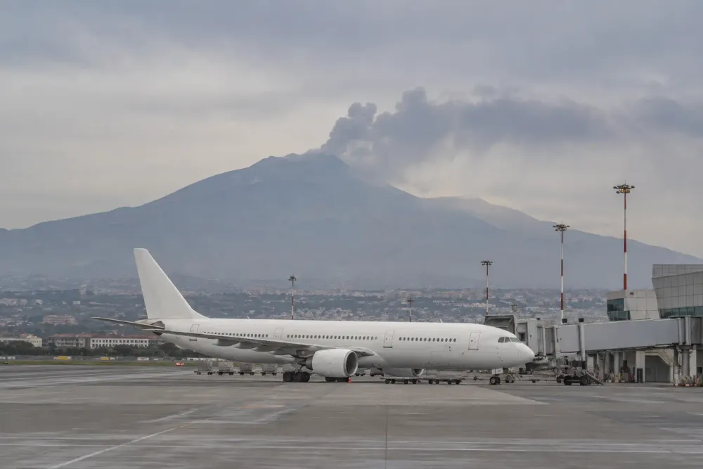 Un avión de pasajeros sin marca visible permanece bajo un cielo brumoso, con el Etna al fondo más allá del aeropuerto.