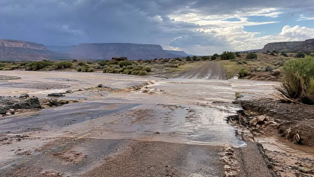 Muddy floodwater crossing a damaged generic desert road beneath clearing storm clouds