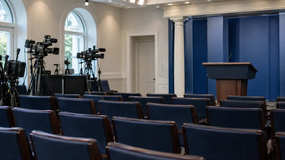 Empty White House-style press briefing room with rows of blue chairs, television cameras and an unbranded lectern