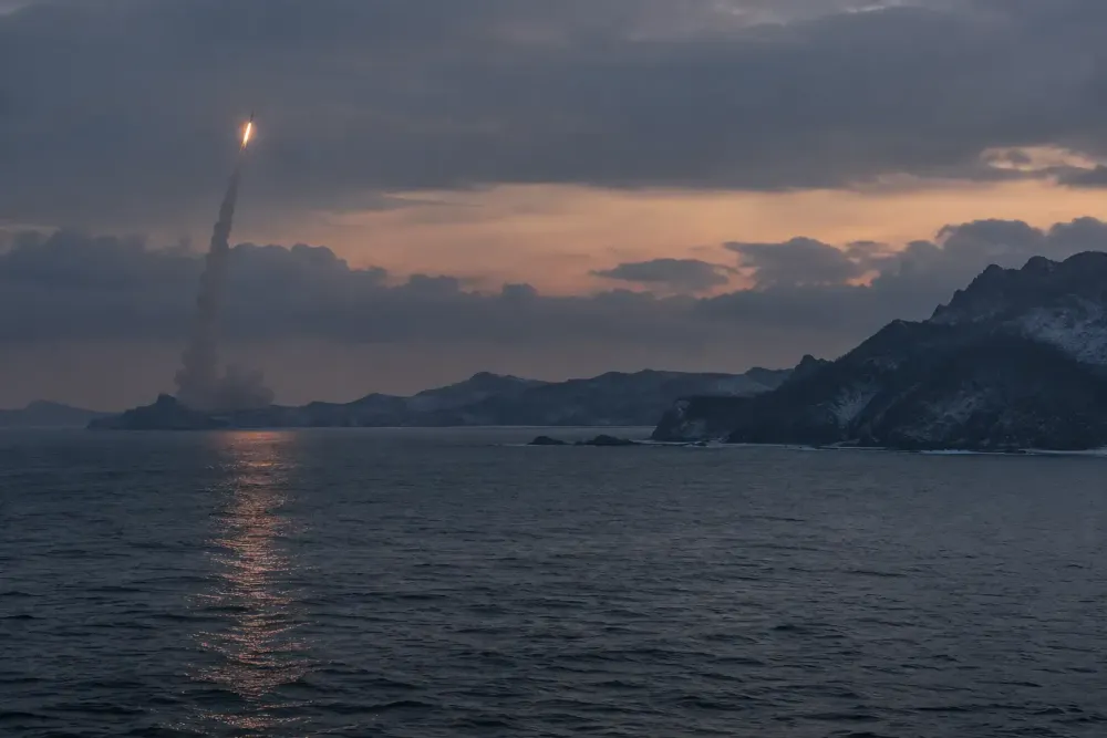 A distant generic missile rises above a rugged coastline at dawn in a restrained editorial scene.