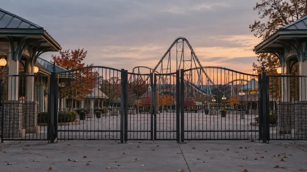 Locked generic amusement park gates with an empty midway and roller coaster silhouette at autumn dusk