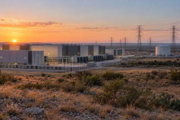A modern data center campus beside transmission lines and water infrastructure in a dry Texas landscape at dawn.
