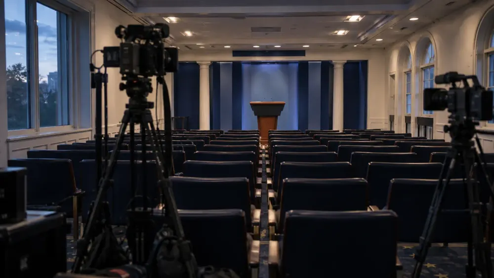 An empty press briefing room with rows of chairs, camera tripods and a neutral podium before dawn.