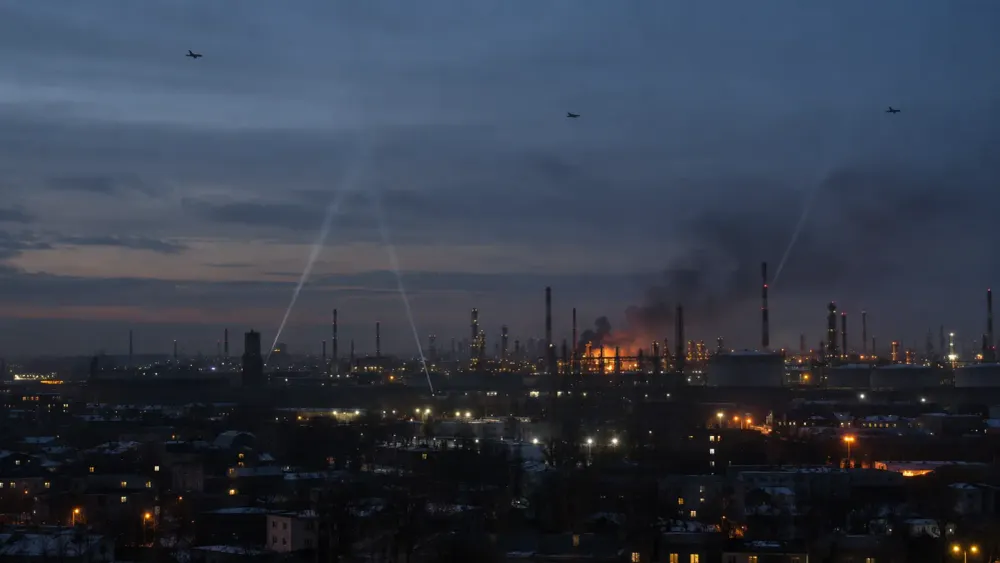 Distant drones and searchlights above an oil refinery at predawn, with smoke rising from an illuminated industrial unit