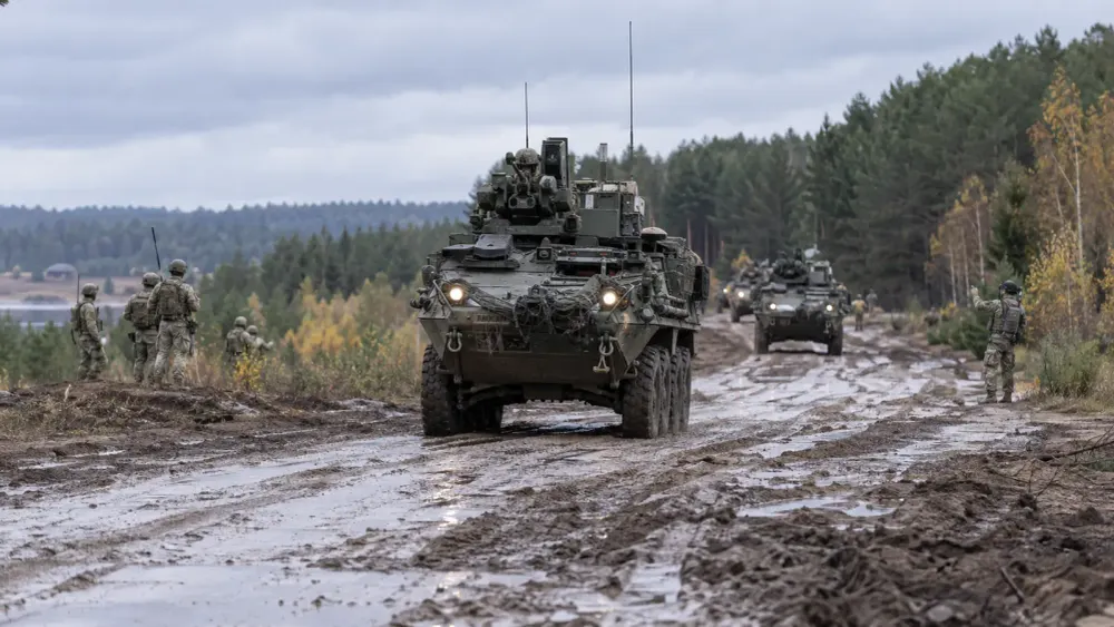 Armored vehicles travel along a muddy training road in a Baltic forest as soldiers coordinate nearby.