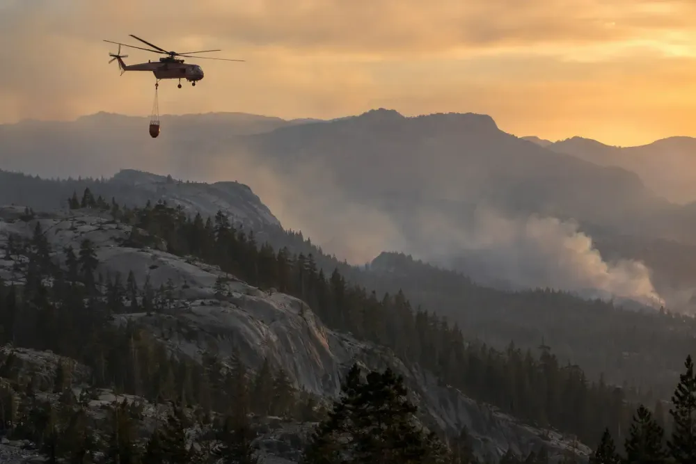 A heavy-lift firefighting helicopter carries a water bucket above a smoke-hazed Sierra Nevada forest.