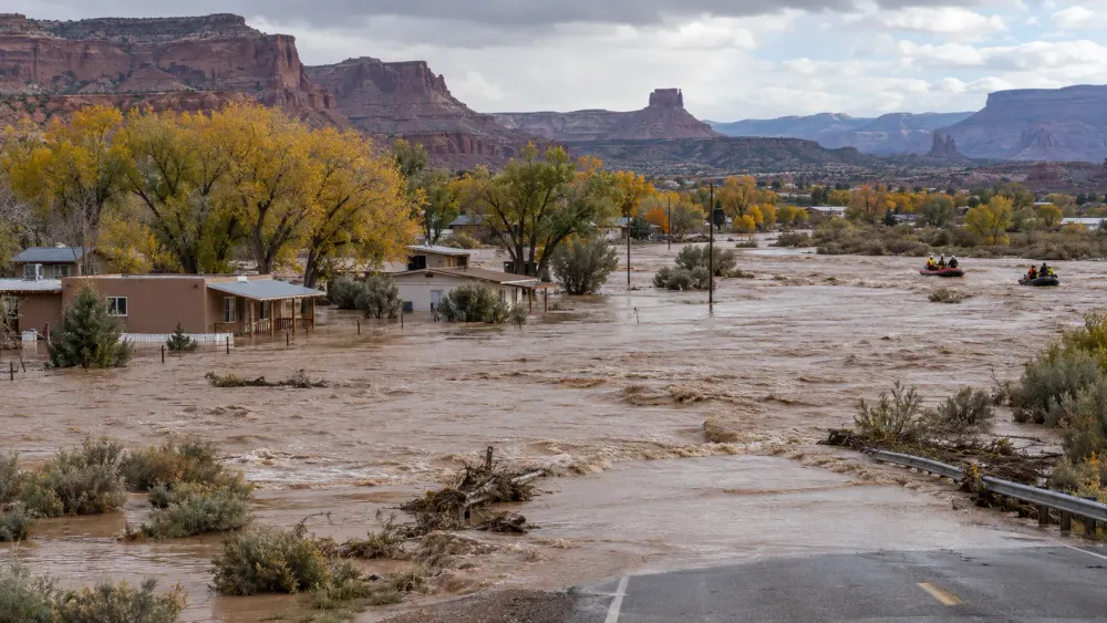 Illustrative view of muddy floodwater surrounding homes and a road in a high-desert community as rescue boats cross the water.