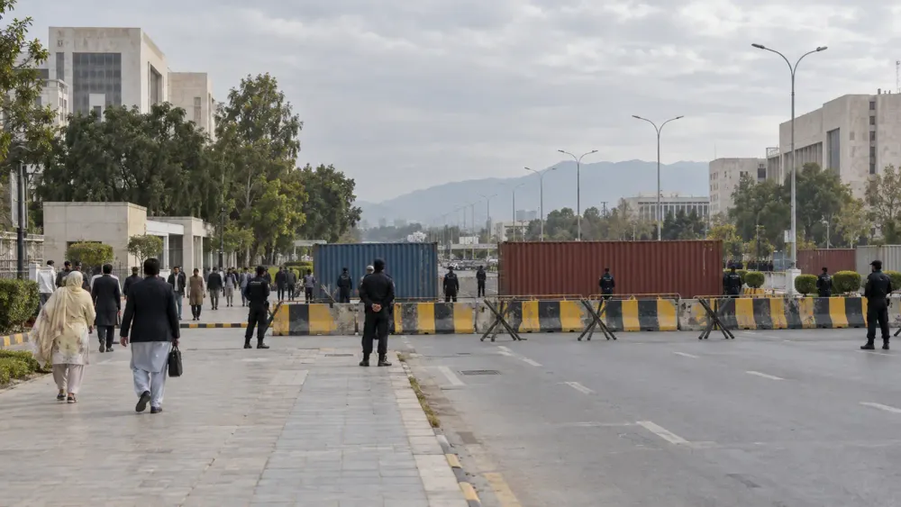 Police officers stand near road barriers outside a generic government complex in Pakistan as civilians move through the area under overcast daylight.