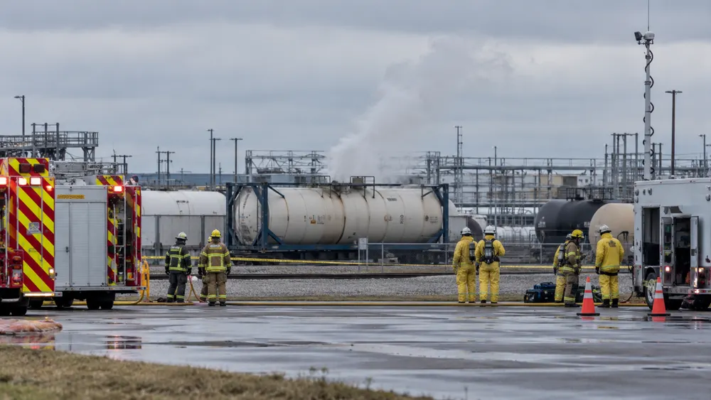 Hazmat responders watch a venting intermodal tank from behind a safety perimeter at a generic industrial transport yard.