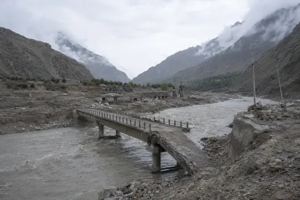 Flood-damaged Himalayan river valley with a broken concrete bridge, mud-covered structures and swollen water under overcast skies.