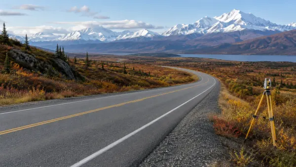 An empty Alaska highway curves through autumn tundra toward snow-covered mountains beside a survey tripod.