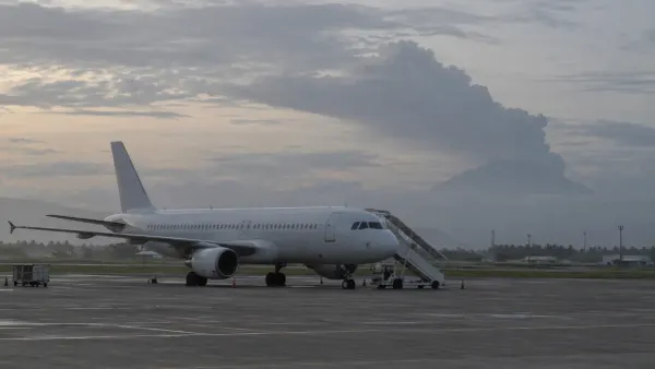 Un avión de pasajeros sin logotipo en la pista de un aeropuerto tropical, con una columna de ceniza volcánica a lo lejos
