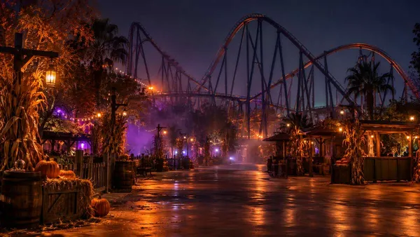 Generic Southern California amusement park midway at night with a roller coaster silhouette, Halloween decorations, amber lights and theatrical fog.