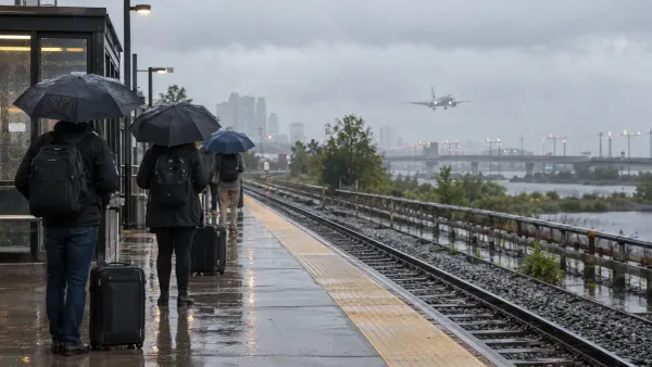 Viajeros con paraguas esperan junto a vías ferroviarias empapadas por la lluvia mientras un avión se aproxima entre la…