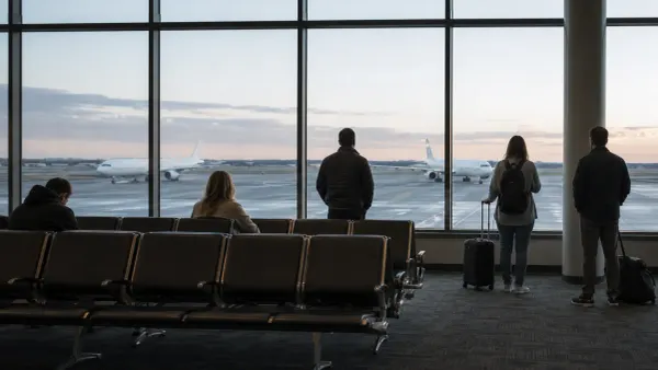 Travelers wait beside empty seats while unbranded passenger aircraft stand outside an international airport terminal