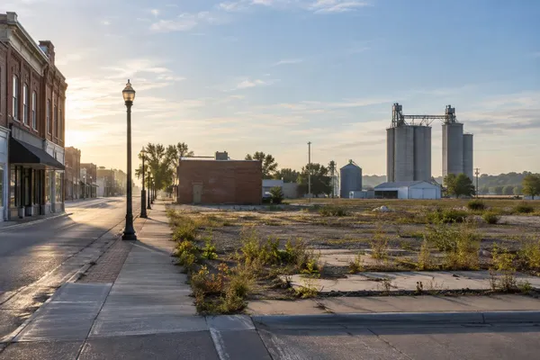 A quiet Illinois commercial corridor beside an underused redevelopment lot and grain elevators at sunrise